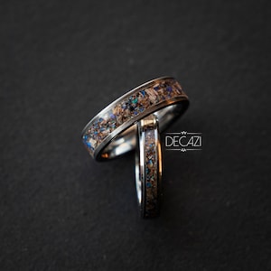 May include: Two silver rings with a unique design featuring embedded blue and brown particles. The rings are stacked on top of each other.