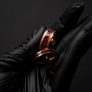 K&ouml;nnte beinhalten: Zwei Ringe aus Ros&eacute;gold mit einer roten Einlage. Die Ringe sind &uuml;bereinander gestapelt und werden in einer schwarzen behandschuhten Hand gehalten.
