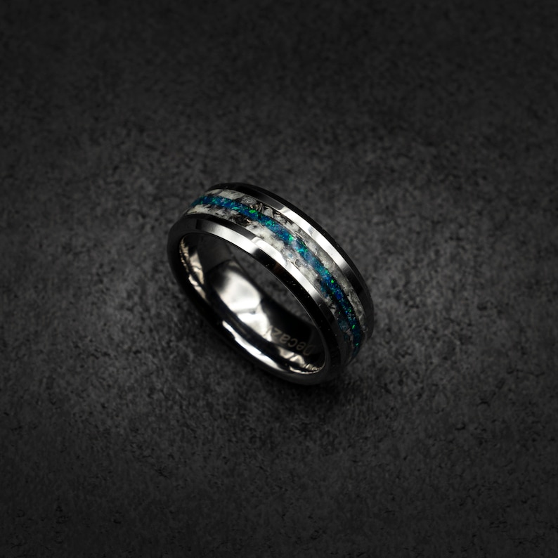 May include: A black tungsten carbide ring with a white and blue inlay. The inlay is made of crushed opal and features a sparkly, iridescent finish.