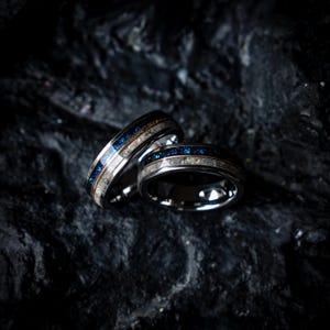 Op de afbeelding: Twee zilveren ringen met een donkerblauwe en witte inleg en een dunne gouden band. De ringen hebben een donkere achtergrond.