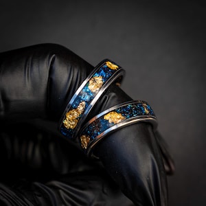 Op de afbeelding: Twee blauwe en gouden ringen met een glinsterende afwerking. De ringen zijn op elkaar gestapeld en worden vastgehouden in een zwarte handschoen.