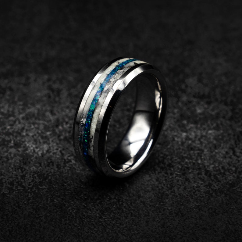 May include: A silver tungsten carbide ring with a blue and white inlay. The ring has a bevelled edge and a polished finish.
