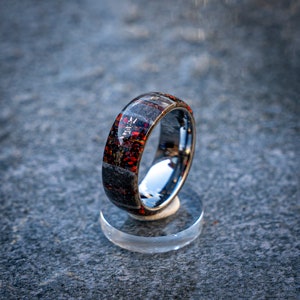 Peut inclure: Une bague en carbure de tungstène noire avec un incrustation rouge et grise. La bague est sur un présentoir transparent.