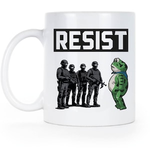 May include: White ceramic mug with the word "RESIST" in bold black letters. The mug features an illustration of four armed figures in black uniforms facing a green frog with a blue scarf.