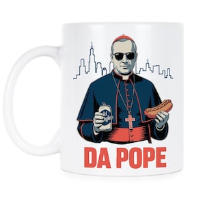 May include: White ceramic mug featuring a cartoon illustration of a figure in religious garb wearing sunglasses, holding a can of beer and a hot dog. The mug has the text "DA POPE" in red block letters, with a city skyline in the background.