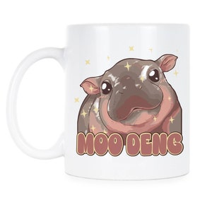 May include: White ceramic mug with a cartoon illustration of a brown and pink hippopotamus with sparkling eyes. The hippopotamus is looking at the viewer and the text "MOO DENG" is printed below the image.