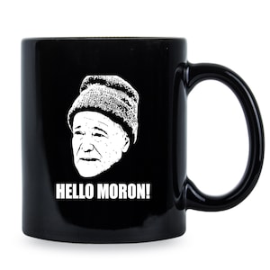 May include: A black ceramic mug featuring a white graphic illustration of a person wearing a knit beanie cap. Below the illustration, the text HELLO MORON! is printed in bold white capital letters. The mug has a curved handle on the right side.