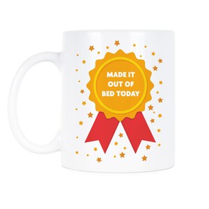 May include: White ceramic coffee mug with a gold and red ribbon design. The ribbon has the text "Made it out of bed today". The background is white with gold stars.