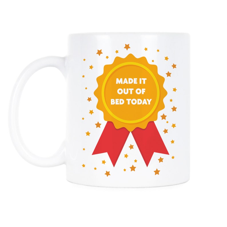 May include: White ceramic coffee mug with a gold and red ribbon design. The ribbon has the text "Made it out of bed today". The background is white with gold stars.