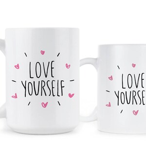 May include: Two white ceramic mugs with black text that reads "LOVE YOURSELF" surrounded by pink hearts.