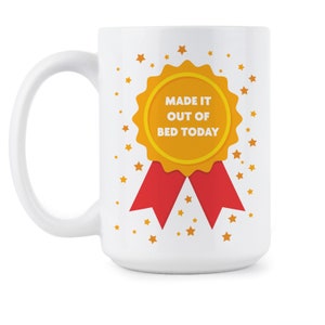May include: White ceramic coffee mug with a gold and red design. The design features a gold medal with the text "Made it out of bed today".