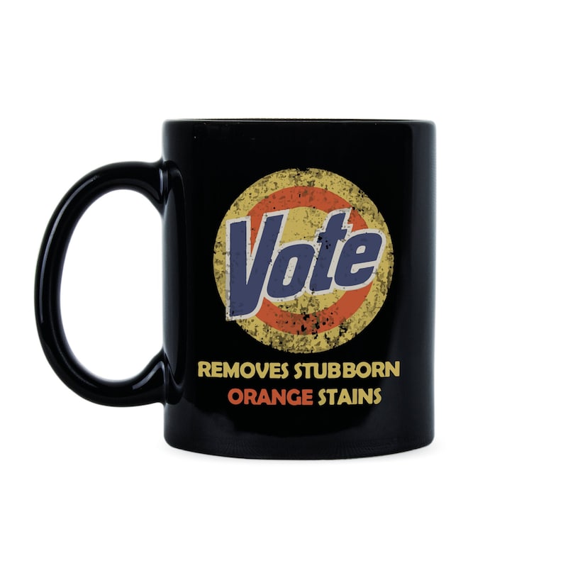 May include: Black coffee mug with a white circle and the word "Vote" in blue letters. The circle has a red and yellow design. The text below the circle reads "Removes Stubborn Orange Stains".