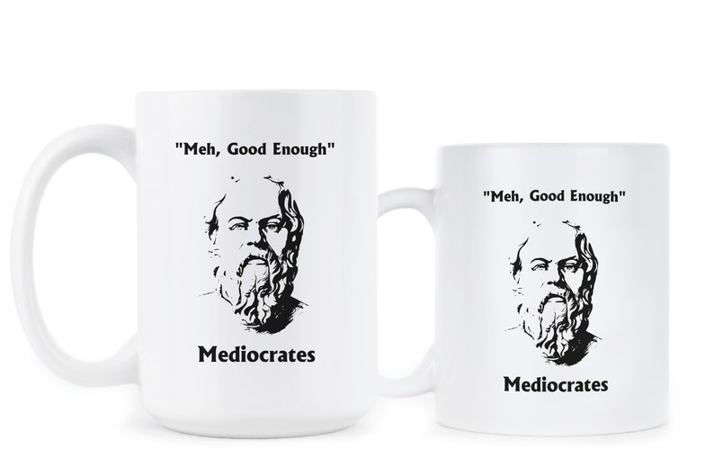 May include: Two white ceramic mugs with black text and a black and white image of a man with a beard. The text on the mugs reads "Meh, Good Enough" and "Mediocrates".