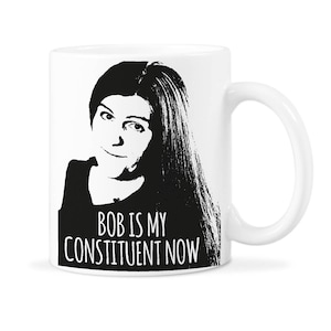 May include: White ceramic mug featuring a black and white image of a woman with long hair and the text "BOB IS MY CONSTITUENT NOW".