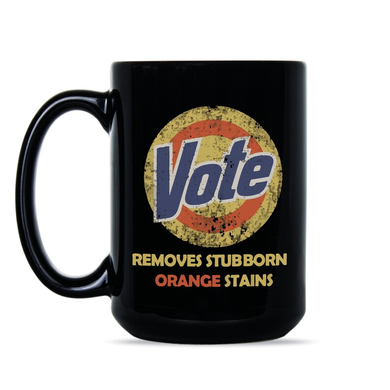 May include: Black coffee mug with a white circle and the word "Vote" in blue text. The circle has a red and yellow design and the text "Removes Stubborn Orange Stains" is below the circle.