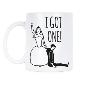 Puede incluir: Taza de café de cerámica blanca con un dibujo de línea negra de una novia arrastrando a un novio del brazo. El texto "I GOT ONE!" está impreso en negro en la taza.