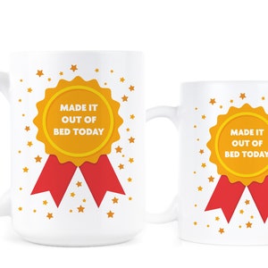 May include: Two white ceramic mugs with a gold and red ribbon design. The mugs feature a gold medal with the text "Made it out of bed today".