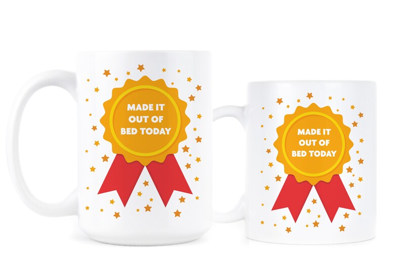 May include: Two white ceramic mugs with a gold and red ribbon design. The mugs feature a gold medal with the text "Made it out of bed today".