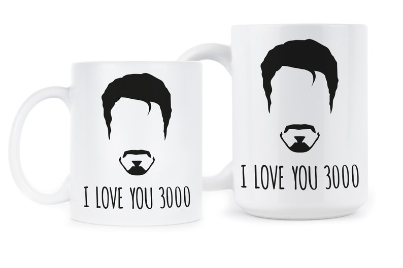 May include: Two white ceramic mugs with black silhouettes of a man's face and the text "I love you 3000".
