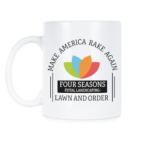 May include: White ceramic coffee mug with a black and white graphic design. The design features a stylized flower with four petals in green, blue, orange, and red. The text reads "Make America Rake Again" and "Four Seasons - Total Landscaping - Lawn and Order".