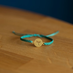 Puede incluir: Una pulsera de macramé turquesa con un dije circular dorado con la silueta de una libélula. La pulsera es ajustable y tiene un diseño trenzado. El fondo es borroso, turquesa y marrón.
