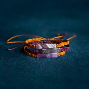 Op de afbeelding: Drie gestapelde armbanden met paarse en witte stenen accenten. De armbanden zijn gemaakt van geweven oranje en paarse koord.