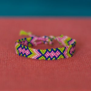 May include: A colourful friendship bracelet with a geometric pattern in pink, green, blue and white.