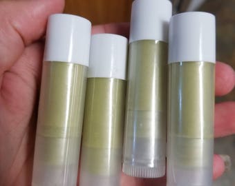 May include: Four lip balm tubes with white caps and clear bases. The balm inside is a light green color. The tubes are held in a hand, and the background is a blurred view of a wooden surface.