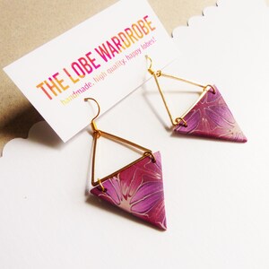 Pode incluir: Um par de brincos triangulares com um padrão floral roxo e rosa, presos a armações de metal dourado com ganchos. Os brincos são apresentados num cartão branco com o texto THE LOBE WARDROBE handmade. high quality. happy lobes!