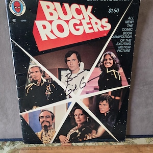 May include: A Buck Rogers Giant Movie Edition comic book featuring a black cover with white stars, the title in red, and multiple character photos. A signature is visible on the cover. A wooden ruler is placed below, showing a 12-inch length.
