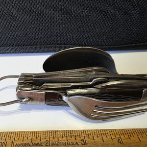 May include: A multi-tool camping utensil set with a spoon, fork, knife, and other tools. The set is made of stainless steel and has a ring for attaching to a keychain. The ruler below the utensil set measures in inches.