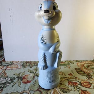 May include: A blue plastic toy bottle shaped like a cartoon squirrel. The bottle has a white face with black eyes and a black nose. The bottle is labeled "THUMPER" at the bottom.