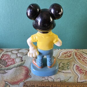 May include: A vintage plastic Mickey Mouse figurine wearing a yellow shirt, blue jeans, and black shoes. The figurine is standing on a blue base with the text "Walt Disney Productions" and "Made in Hong Kong".