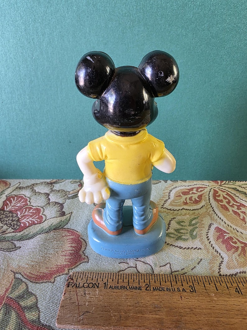May include: A vintage plastic Mickey Mouse figurine wearing a yellow shirt, blue jeans, and black shoes. The figurine is standing on a blue base with the text "Walt Disney Productions" and "Made in Hong Kong".