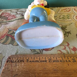 May include: A vintage plastic figurine of a cartoon dog wearing a yellow shirt, blue pants, and a red bow tie. The figurine is standing on a blue base.