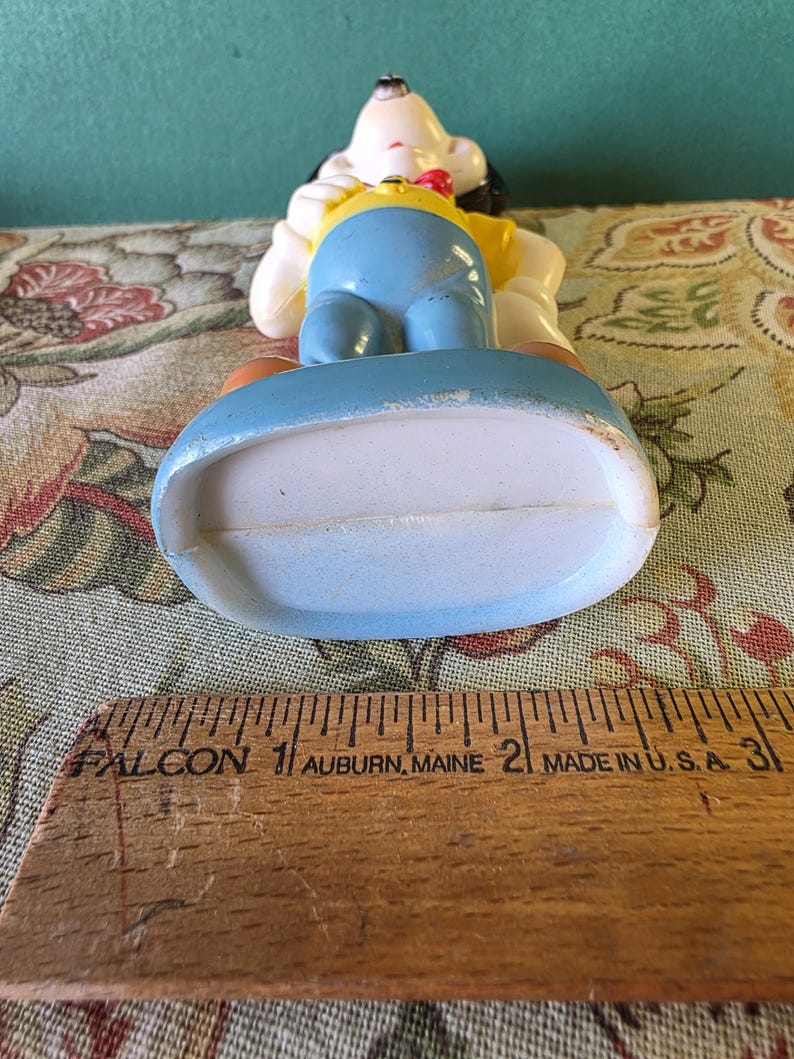 May include: A vintage plastic figurine of a cartoon dog wearing a yellow shirt, blue pants, and a red bow tie. The figurine is standing on a blue base.