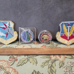 May include: Two embroidered military patches and two circular challenge coins are displayed alongside a wooden ruler. The left patch features a lightning bolt and the text Strategic Air Command. The right patch shows a sword and wings with Tactical Air Command.