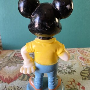May include: A plastic figurine of Mickey Mouse wearing a yellow shirt and blue jeans. The figurine is standing on a blue base with the text "Walt Disney Productions Hong Kong" printed on it.