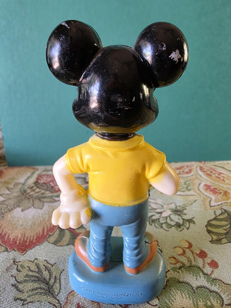 May include: A plastic figurine of Mickey Mouse wearing a yellow shirt and blue jeans. The figurine is standing on a blue base with the text "Walt Disney Productions Hong Kong" printed on it.