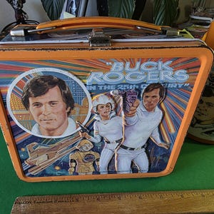 May include: Vintage orange metal lunchbox featuring Buck Rogers in the 25th Century. The lunchbox has a portrait of Buck Rogers and other characters. A matching orange thermos is also visible.