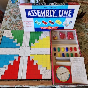 May include: Vintage board game titled "Assembly Line." The box is white with blue and red text and illustrations of cars. The game includes a colorful game board, miniature car models, and game components. The game is designed for 2-4 players.