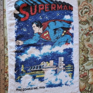 May include: A white towel with a blue and red Superman image. The image shows Superman flying through the air with a city skyline in the background. The text "SUPERMAN" is printed in red letters at the top of the towel. The text "SDC COMICS INC. 1980" is printed in black letters at the bottom of the towel.