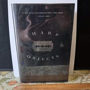 Puede incluir: Un libro de tapa dura negro titulado "Sharp Objects" de Gillian Flynn. La portada presenta texto blanco y un gráfico de cuchilla de afeitar. Incluye una cita de Harlan Coben.