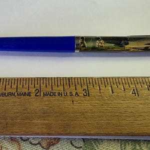 May include: A blue pen with a clear barrel featuring a scenic design. A wooden ruler with black markings and the text "FALCON AUBURN, MAINE MADE IN U.S.A." is below the pen.