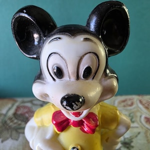 May include: A vintage plastic Mickey Mouse figurine with black ears, a white face, and a yellow shirt with a red bow tie.