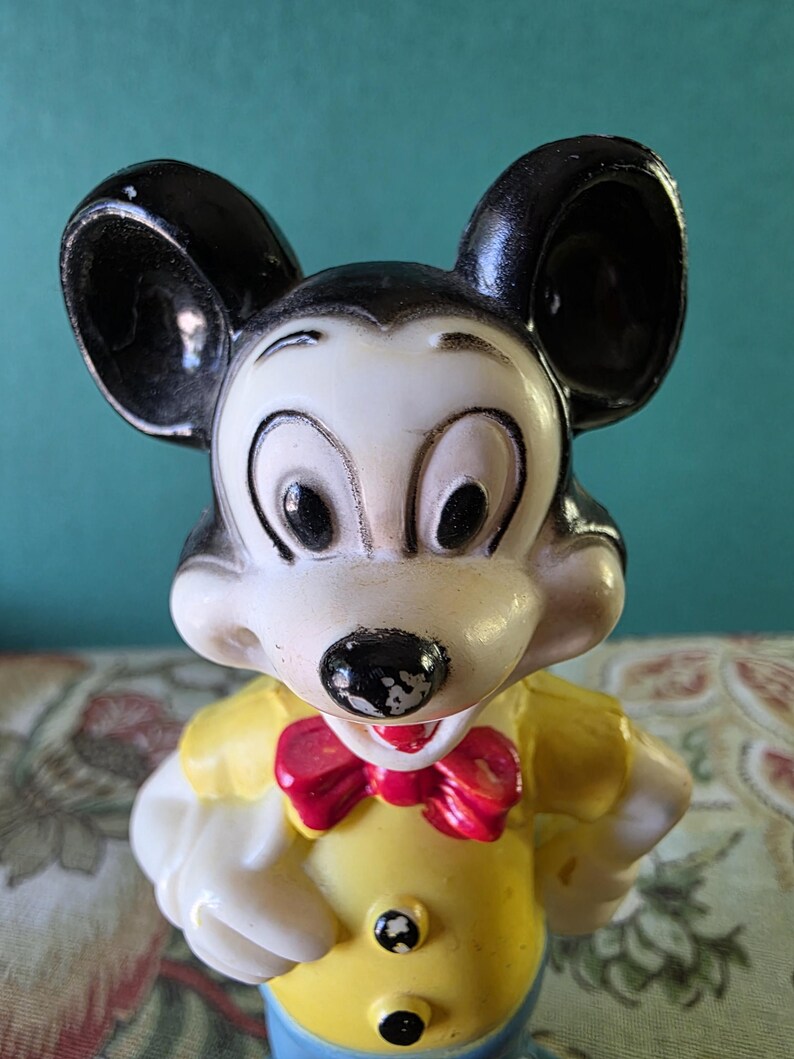 May include: A vintage plastic Mickey Mouse figurine with black ears, a white face, and a yellow shirt with a red bow tie.