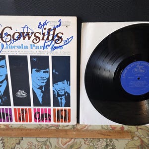 May include: An album cover for "The Cowsills plus the Lincoln Park" with signatures. The cover features a design with the band members in blue and black, with a black vinyl record next to it. The record has a blue label.