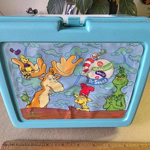 May include: A light blue plastic lunchbox featuring characters from "The Wubbulous World of Dr. Seuss." The front panel displays illustrations of the Grinch, Horton, and other Seuss characters. The lunchbox has a blue handle and a secure latch.