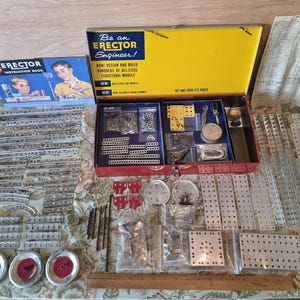 May include: An Erector Set with various metal construction pieces, including beams, plates, wheels, and screws. The set includes an instruction book and a box with the text "Be an Erector Engineer!" in yellow and red.