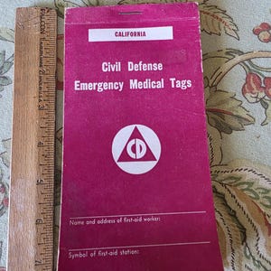 May include: A vintage magenta Civil Defense Emergency Medical Tag from California. The tag features white text and a circular logo with a triangle and a 'CD' symbol. A wooden ruler is next to the tag.